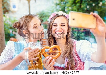 Best friends in Bavarian Tracht making a Selfie with the phone, beer, and a pretzel