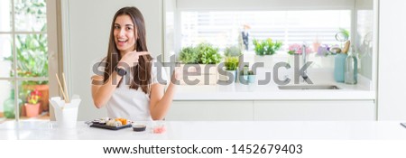Wide angle picture of beautiful young woman eating asian sushi from delivery amazed and smiling to the camera while presenting with hand and pointing with finger.