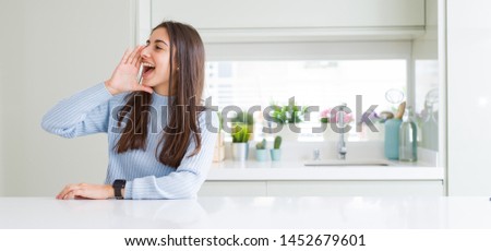 Wide angle picture of beautiful young woman sitting on white table at home shouting and screaming loud to side with hand on mouth. Communication concept.