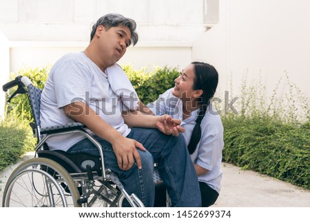 The wife is taking care of the husband sitting on wheelchair,  who is suffering from neurological diseases, or hemiplegia having a facial palsy and kinking fingers, to health and paralysis concept. Royalty-Free Stock Photo #1452699374