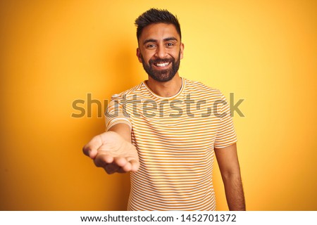 Young indian man wearing t-shirt standing over isolated yellow background smiling cheerful offering palm hand giving assistance and acceptance.