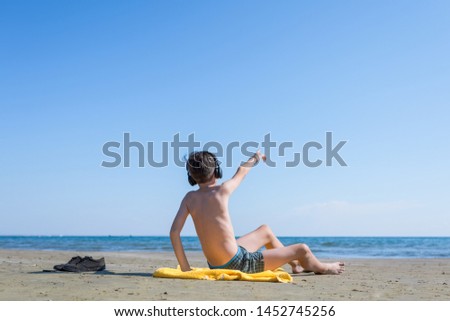 Teen  boy is sitting on yellow towel in the headphones and sunbathes on the beach on the sea and sky background. Concept