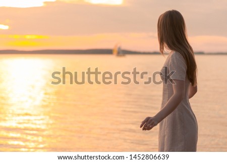 the girl, in a neat delicate cream dress, turned toward the sea. Very beautiful hand bend. The sea at sunset On the horizon from a distance is a sailboat. Cloudy sky Royalty-Free Stock Photo #1452806639