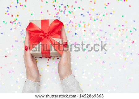 White desk with scattered tinsel, woman holds gift, woman's hands, gift box with red ribbon, close up Royalty-Free Stock Photo #1452869363