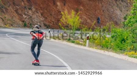 Tall athlete riding his longboard fast and steady downhill while wearing red t-shirt and black jeans.