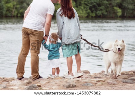 A woman, a man and a little girl are standing near by a lake with their dog. White samoyed dog. Happy family concept. Family day.