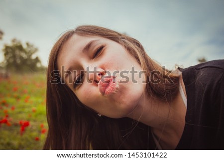 Young woman making duckface kiss while taking selfie picture with her smartphone or camera in field of red poppies. She having fun doing silly and funny to the camera.