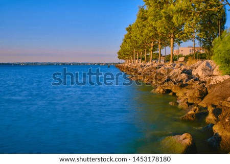 Sunrise on Lake Garda, reflections of trees in the water. The coastline of stones. Photo taken on a long exposure.