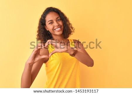 Young brazilian woman wearing t-shirt standing over isolated yellow background smiling in love showing heart symbol and shape with hands. Romantic concept.
