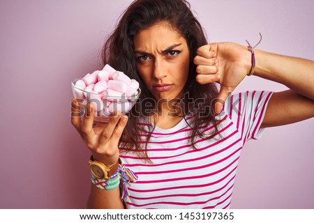 Young beautiful woman holding bowl with marshmallows over isolated pink background with angry face, negative sign showing dislike with thumbs down, rejection concept
