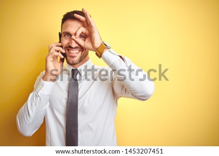 Young handsome business man talking on the phone over yellow isolated background with happy face smiling doing ok sign with hand on eye looking through fingers