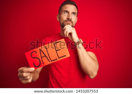 Young man holding sale advertising poster board over red isolated background serious face thinking about question, very confused idea