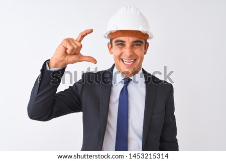 Young handsome architect man wearing suit and helmet over isolated white background smiling and confident gesturing with hand doing small size sign with fingers looking and the camera.