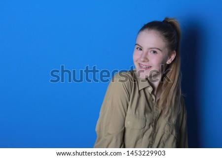 Happy joyful student with braces on teeth posing over blue studio background. Blonde teenage girl with horsetail hairstyle looking and smiling at camera. Brackets, dental service