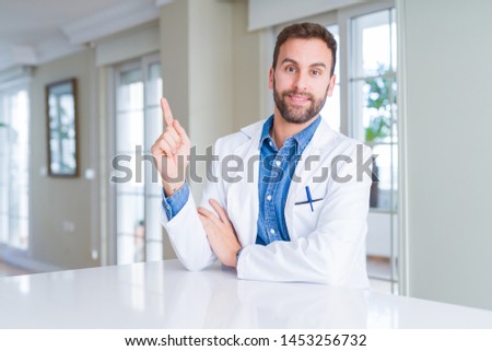 Handsome doctor man wearing medical coat at the clinic with a big smile on face, pointing with hand and finger to the side looking at the camera.