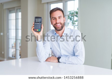 Handsome young man holding point of sale terminal with a happy face standing and smiling with a confident smile showing teeth