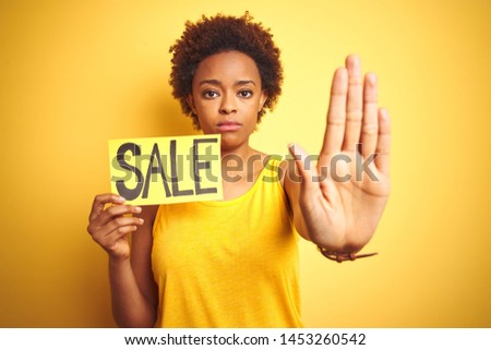 African american woman holding sale board over yellow isolated background with open hand doing stop sign with serious and confident expression, defense gesture