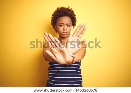 Beauitul african american woman wearing summer t-shirt over isolated yellow background Rejection expression crossing arms and palms doing negative sign, angry face