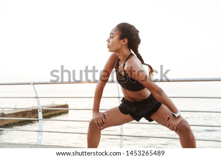Image of focused young woman in sportive clothes stretching her body while doing workout by seaside in morning