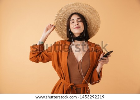 Beautiful young woman wearing straw hat standing isolated over beige background, listening to music with earphones, holding mobile phone