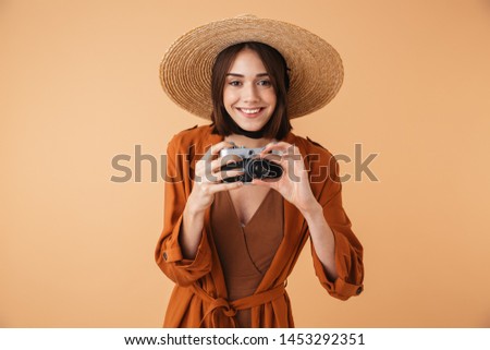 Beautiful young woman wearing straw hat and summer outfit standing isolated over beige background, taking pictures with photo camera
