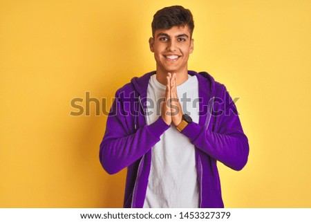 Young indian man wearing purple sweatshirt standing over isolated yellow background praying with hands together asking for forgiveness smiling confident.