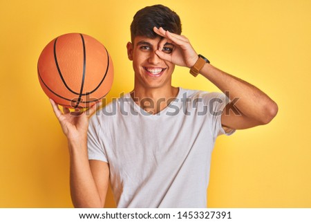 Young indian sportsman holding basketball ball standing over isolated yellow background with happy face smiling doing ok sign with hand on eye looking through fingers