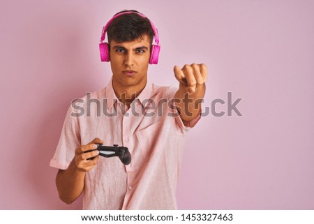 Indian gamer man playing video game using headphones over isolated pink background pointing with finger to the camera and to you, hand sign, positive and confident gesture from the front