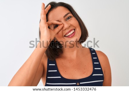 Young beautiful woman wearing blue striped t-shirt standing over isolated white background with happy face smiling doing ok sign with hand on eye looking through fingers