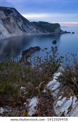 The sun is rising in the horizon, painting the sky pink. Below a trail of rocks enhancing the scene. Wild flowers and grass in the foreground pop a little green into the image.