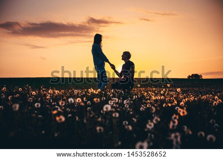 Silhouettes of a man making marriage proposal to his girlfriend on the spring meadow at sunset. Landscape with silhouette of lovers against colorful sky. Couple. People, relationship.