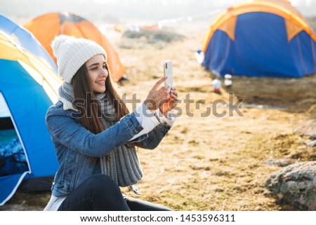 Cheerful young girl sitting at the campsite outdoors, using mobile phone, taking a selfie