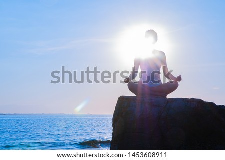 Woman is meditating on the calm beach at sunset. Woman is practicing yoga sitting on stone in Lotus pose at sunset. Silhouette of woman meditating on the beach