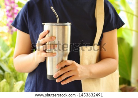 Young woman show reusable glasses of water in her hand with green garden background.Use water-glass and steel straw for Replacement plastic cup can save the earth.Reduce global warming concept. Royalty-Free Stock Photo #1453632491