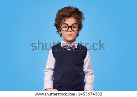 Adorable kid in nerdy glasses and school uniform looking at camera while standing against blue background