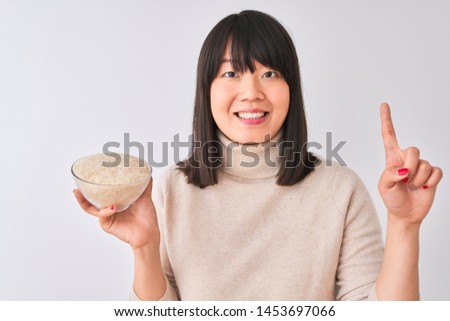 Young beautiful Chinese woman holding bowl with rice over isolated white background surprised with an idea or question pointing finger with happy face, number one