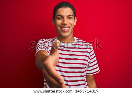 Young handsome arab man wearing striped t-shirt over isolated red background smiling friendly offering handshake as greeting and welcoming. Successful business.