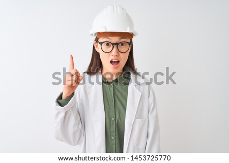 Young chinese engineer woman wearing coat helmet glasses over isolated white background pointing finger up with successful idea. Exited and happy. Number one.