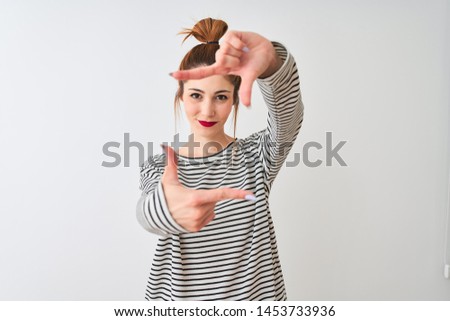 Redhead woman wearing navy striped t-shirt standing over isolated white background smiling making frame with hands and fingers with happy face. Creativity and photography concept.