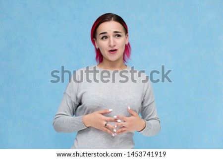Portrait to the waist of a pretty girl with red hair on a blue background in a gray sweater. Standing in the studio right in front of the camera with emotions, talking, showing hands, smiling