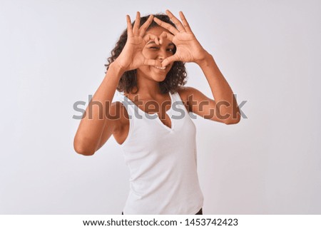 Young brazilian woman wearing casual t-shirt standing over isolated white background Doing heart shape with hand and fingers smiling looking through sign