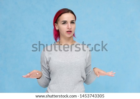 Portrait to the waist of a pretty girl with red hair on a blue background in a gray sweater. Standing in the studio right in front of the camera with emotions, talking, showing hands, smiling