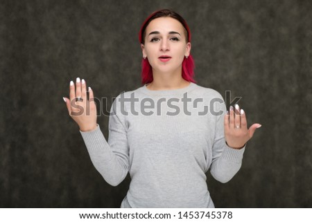Portrait to the waist of a pretty girl with red hair on a gray background in a gray sweater. Standing right in front of the camera in a studio with emotions, talking, showing hands, smiling