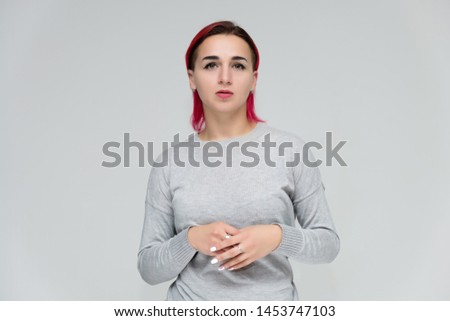 Portrait to the waist of a pretty girl with red hair on a white background in a gray sweater. Standing right in front of the camera in a studio with emotions, talking, showing hands, smiling