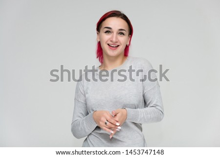 Portrait to the waist of a pretty girl with red hair on a white background in a gray sweater. Standing right in front of the camera in a studio with emotions, talking, showing hands, smiling