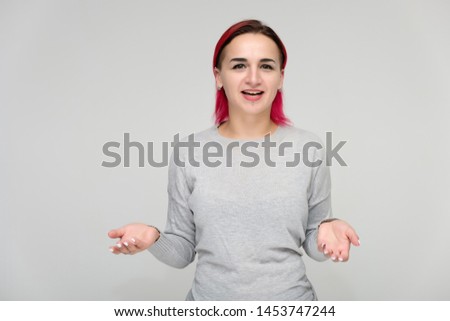 Portrait to the waist of a pretty girl with red hair on a white background in a gray sweater. Standing right in front of the camera in a studio with emotions, talking, showing hands, smiling