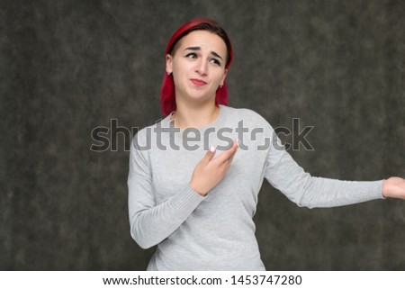 Portrait to the waist of a pretty girl with red hair on a gray background in a gray sweater. Standing right in front of the camera in a studio with emotions, talking, showing hands, smiling