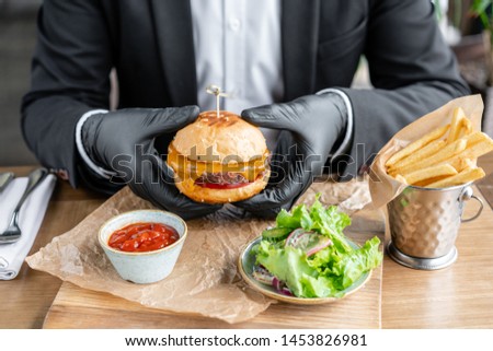 Young man in business suit and black gloves eating delicious juicy cheeseburger with beef chop. Burgers in the cafe with salad and fries, fast food.