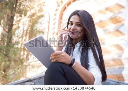 portrait of a beautiful indian girl. Business woman, wearing glasses, smiling, sitting on the steps of an office building, holding a tablet in her hand