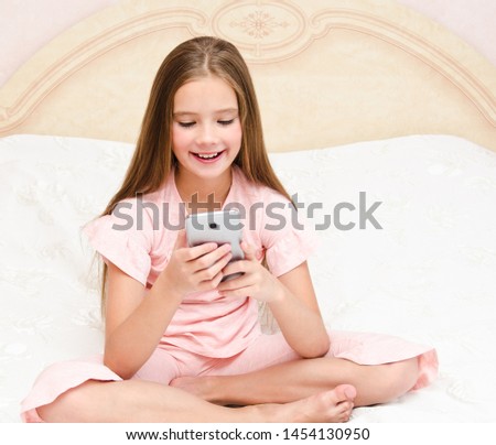 Portrait of cute smiling  little girl child playing with cell phone smartphone sitting no the bed at home